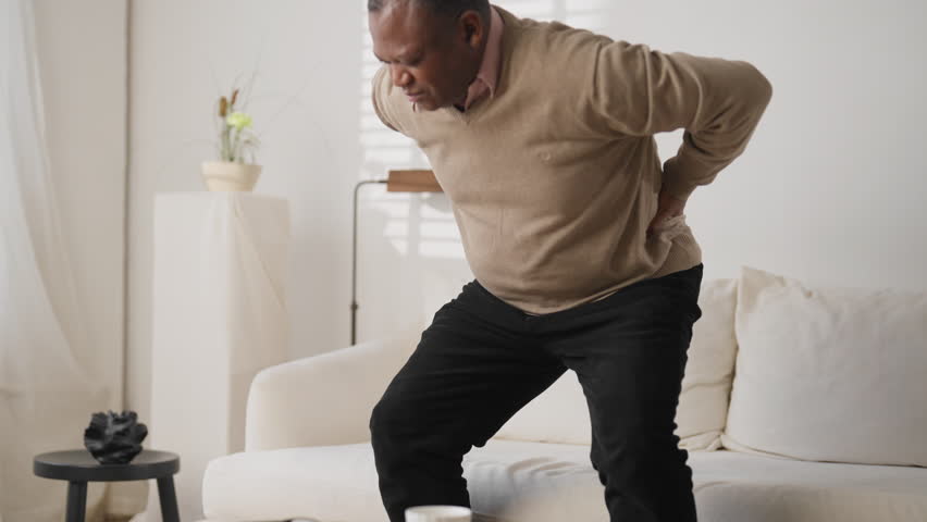 Senior black man feeling lower back pain, radiculitis, disease of old age. African american male person sitting on couch in living room and suffering of backache, aging and healthcare, stay active