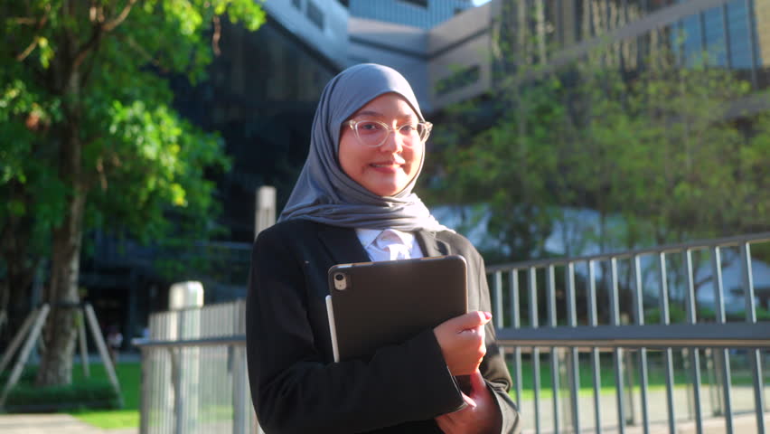 Portrait of a confident Asian Muslim businesswoman wearing hijab and glasses, holding a tablet and smiling outdoors against a modern office building and green trees background.