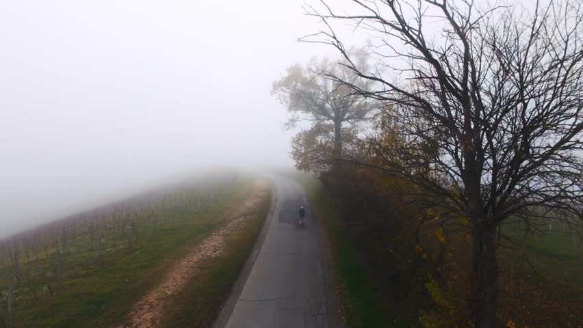 Aerial view of cyclist riding through misty rural vineyard road surrounded by farmland in foggy weather.