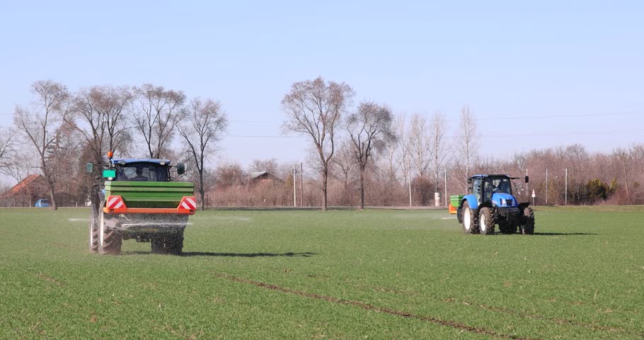 Two tractors with spreader fertilizer working on wheat field, slow motion. Farmers with agricultural machinery fertilizing wheat field in winter time, in a background car and trees. Spreading mineral fertilizer, landscape rural scene