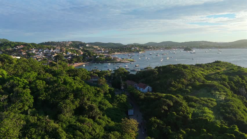 Vegetated hills, beach, and residential area at Praia dos Ossos, Brazil, with multiple boats docked in the ocean during sunset. Forward wide-angle glide.