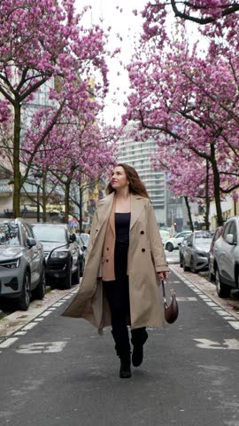 Confident and happy young woman walking down a beautiful city street in spring, enjoying the sight of blooming pink cherry blossom trees lining the road on a lovely day