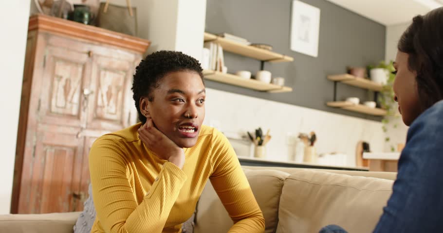 Adult Black woman listening then sharing while Indian friend nodding on beige sofa in living room. Conversational, friendship, warmth, intimacy, modern, cozy, social