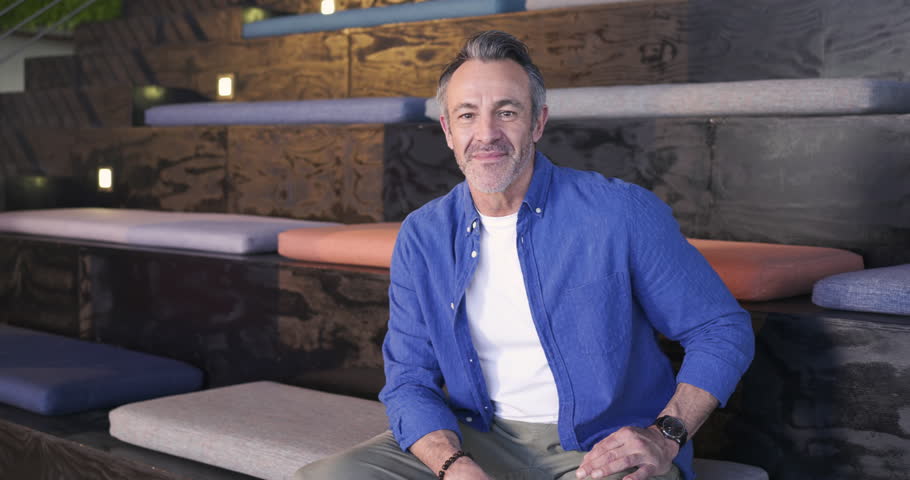 Middle-aged man wearing shirt sitting in modern lounge on bench speaking laughing responding to cue. Relaxation, casual, warmth, conversational, friendliness, comfort, interior