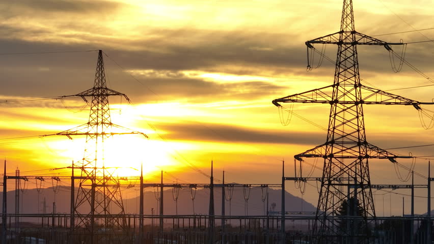 Power lines and electrical transmission towers stand tall in the rural area as the sun sets behind them. The sky shows vibrant colors.