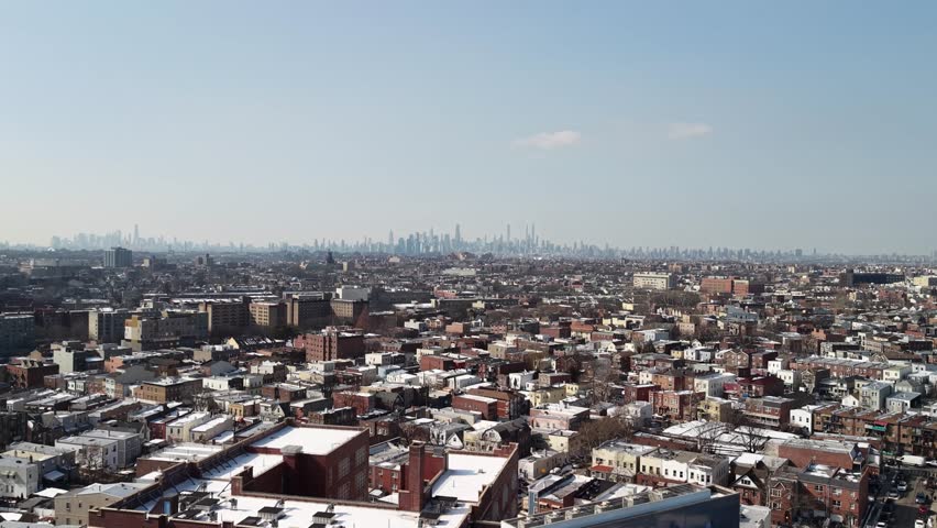 Sweeping aerial view flying over the snow covered rooftops of brooklyn residential neighborhoods with the iconic manhattan skyline visible in the hazy distance during a clear winter day