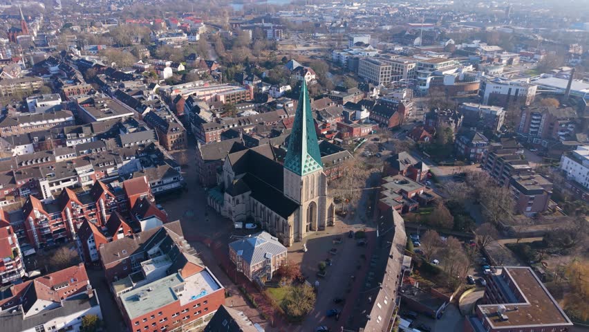 Aerial view of a historic European city center with a prominent Gothic cathedral featuring a green spire, surrounded by traditional red brick buildings and urban landscape in soft daylight.