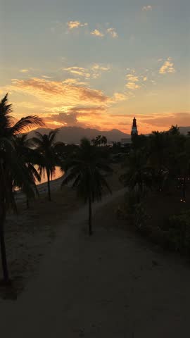 Golden sky fades over palm-lined shore. Silhouettes stand quiet against fading light. Warm hues paint distant mountains and calm waters. Peaceful moment captured by ocean breeze and gentle waves