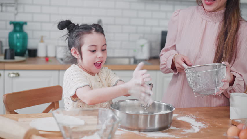 Excited young Asian girl laughs and plays with white flour in a bowl while baking with her mother in a bright, modern kitchen during a fun family morning.
