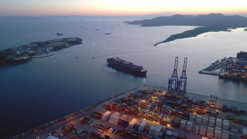 Aerial drone footage of a fully loaded container ship leaving an industrial cargo port during blue hour. Maritime freight transport vessel departing a busy container terminal with logistics infrastructure and shipping operations. Filmed near Athens, Greece.