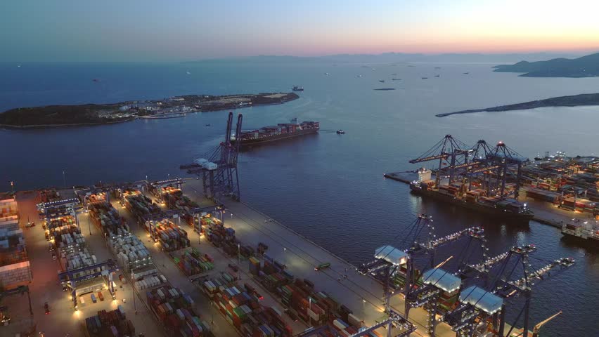 Aerial drone footage of a fully loaded container ship leaving an industrial cargo port during blue hour. Maritime freight transport vessel departing a busy container terminal with logistics infrastructure and shipping operations. Filmed near Athens, Greece.