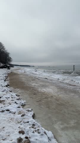 Wide view of frozen Baltic Sea coast with snow covered beach and unusual ice formations rising from the water under a heavy gray winter sky.