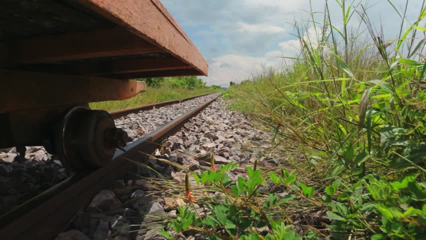 Low-angle cinematic view from beneath the traditional Bamboo Train (Norry) in Battambang, Cambodia. The small rail wheel rolls along a rural railway track surrounded by lush green vegetation