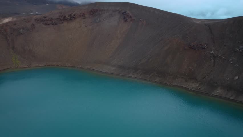 Aerial drone pull out revealing turquoise Viti crater lake surrounded by steep volcanic slopes with calm water surface inside natural caldera Iceland landscape.