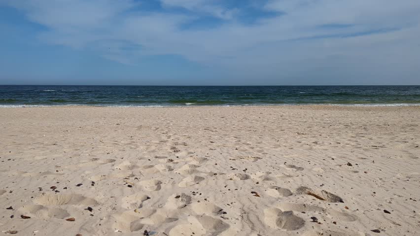 Cinematic small sea waves washing over an empty sandy beach under clear blue skies. Wide open shoreline with no people