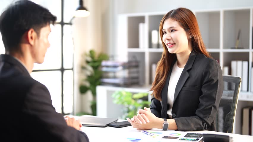 Young Asian businesswoman sitting at her desk, gesturing and smiling while talking to a male candidate during a job interview or a business meeting in a modern, bright corporate office