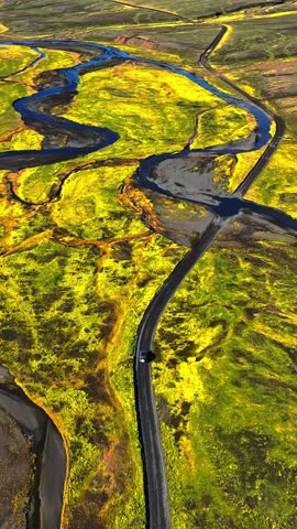 Vertical drone view of a car driving a winding road through bright moss-covered volcanic plains and braided rivers in Iceland, creating a bold cinematic travel landscape.
