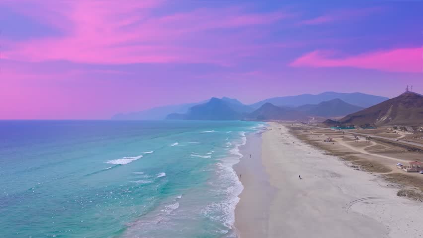 Aerial view of turquoise ocean waves breaking on Socotra a sandy beach alongside mountains under a purple sunset sky in Yemen.