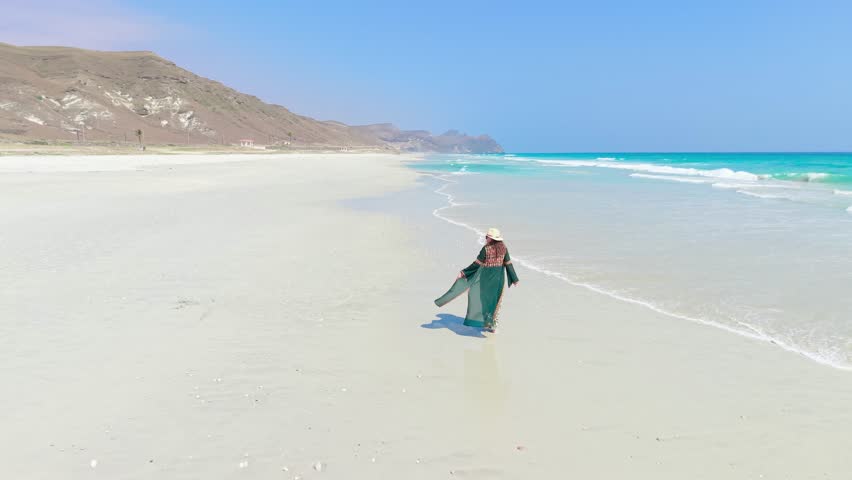 A young woman wearing a green dress and hat walks along the white sand shore beside turquoise ocean water in Socotra, Yemen.