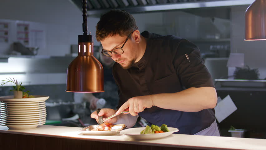 Male chef carefully arranging gourmet duck leg and vegetables on plate with kitchen tweezers under heat lamp while preparing dish in restaurant kitchen
