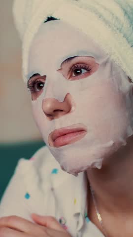 Portrait, beautiful woman in a cosmetic fabric mask and with a white towel on her head, under light from the TV screen. She watches TV at night, lying on a sofa, at home