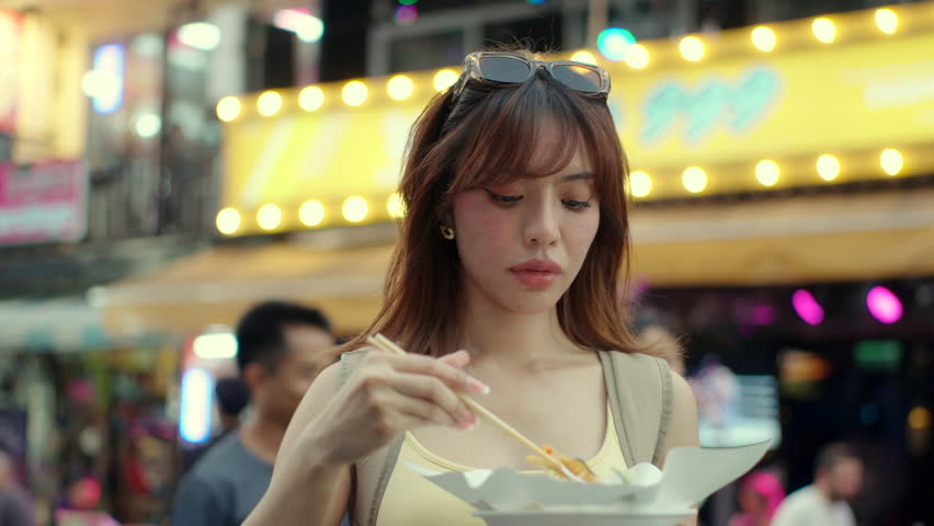 Beautiful trendy woman traveler enjoy eating street food local noodles at night market with nightlife neon atmosphere background, female tourist enjoy local asian cuisine experience.
