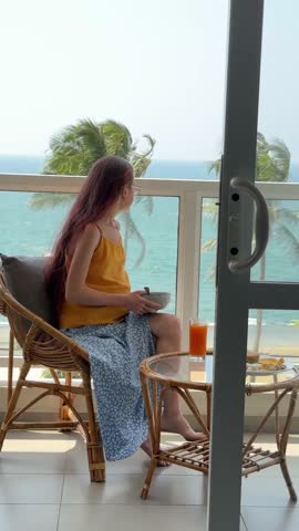 Caucasian woman seated on balcony scrolling phone with breakfast and juice, palm trees bending in wind, digital nomad checking