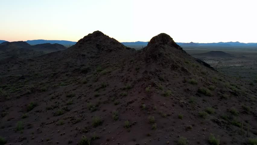 A drone captures a dramatic aerial view as it flies between two dark desert peaks, revealing a long irrigation canal stretching across the vast, arid valley floor at twilight. The scene showcases the rugged beauty of the desert landscape and the engineering of water management.