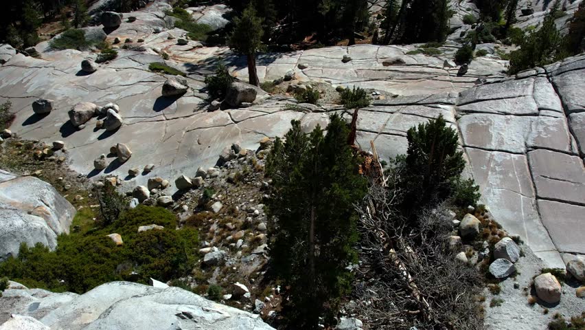 An aerial perspective captures the rugged beauty of Yosemite National Park. Starting low near textured granite rocks and evergreen trees, the camera rises to show the vast, sun-drenched mountain range, including iconic peaks and deep valleys under a brilliant clear blue sky.