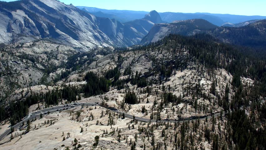 This breathtaking aerial drone shot showcases the vast granite landscape of Yosemite National Park. A winding mountain road snakes through pine-dotted rocky slopes, leading the eye toward the distant iconic Half Dome under a clear blue sky. Perfect for nature and travel projects.