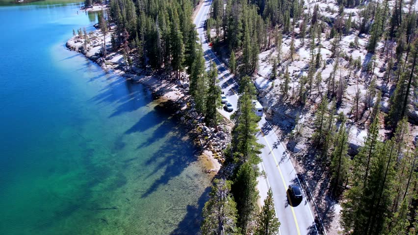 A stunning aerial drone perspective of Tenaya Lake in Yosemite National Park. The video captures the deep blue waters, the winding Tioga Road with passing vehicles, dense pine forests, and the surrounding majestic granite mountains under a clear sky.