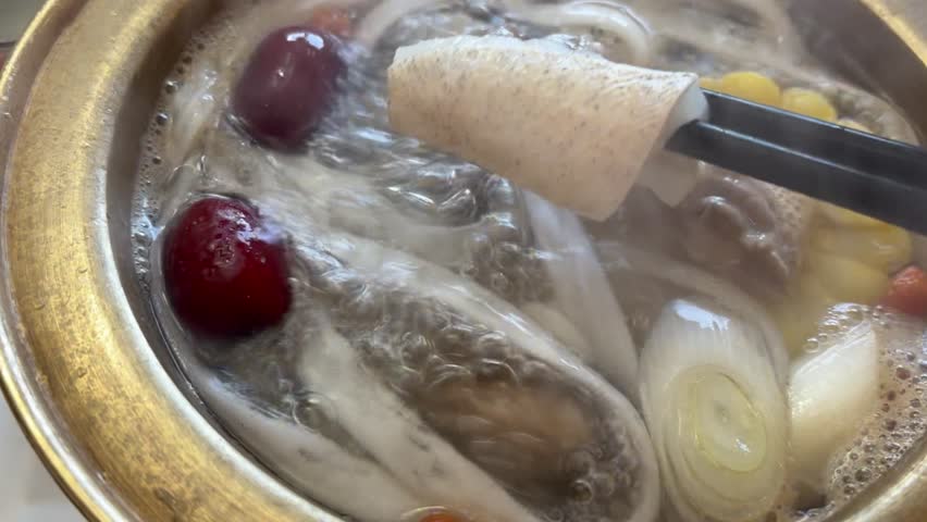 4K close up of a fresh fish slice being placed into a traditional copper hot pot. Visible ingredients: goji berries (Gouqi), radish, scallions, red dates, beef slice, glass noodles (fenshi), and corn.