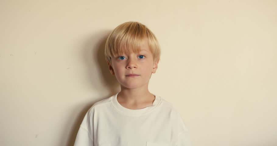 A blond, blue eyed boy aged 5 or 6 calmly and thoughtfully looking into the camera against a light neutral background.