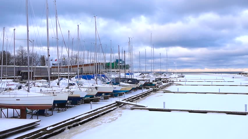 Snow covered sailboats resting on a frozen marina in Pirita with icy harbor and Baltic Sea under winter clouds.