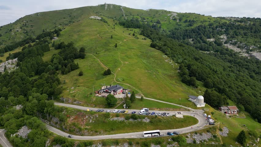 High-angle aerial view of a winding mountain road and parking area next to a traditional alpine hut and observatory on the green slopes of Mount Matajur under a soft, overcast summer sky.