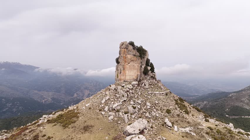 Drone aerial view of Perda Liana, iconic limestone monolith rising above the rugged mountains of Ogliastra, Sardinia, Italy, surrounded by Mediterranean scrub under a cloudy sky