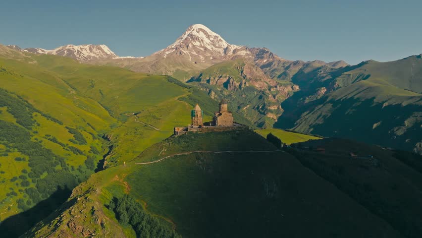Drone view of ancient Gergeti Trinity Church sits atop a green hill, with majestic snow-capped Mount Kazbek (Kazbegi) rising in the background near Stepantsminda, Georgia