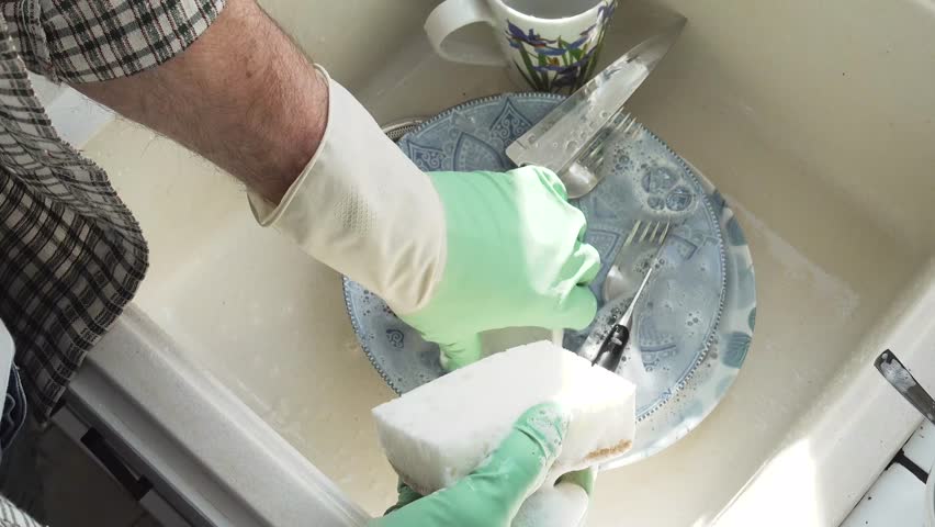 Man in green rubber gloves washes dishes in a kitchen sink using a sponge and liquid soap. Contemporary domestic scene and modern masculinity. Concept of sharing household chores. Day light, close-up