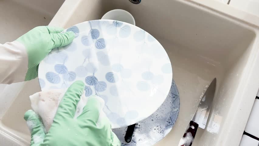 Man in green rubber gloves washes dishes in a kitchen sink using a sponge and liquid soap. Contemporary domestic scene and modern masculinity. Concept of sharing household chores. Day light, close-up