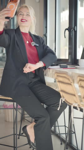 Stylish blonde businesswoman in black suit smiling while taking selfie with smartphone, sit on high chair at modern cafe bar counter during coffee break