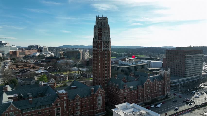 Vanderbilt University in Nashville, with iconic campus tower, surrounding academic buildings, and nearby city streets under bright sunlight. Aerial approaching shot.