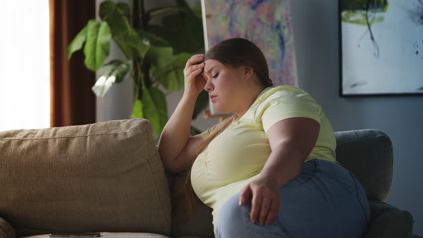 Sad large-bodied woman thinking about problems while sitting on sofa, depression