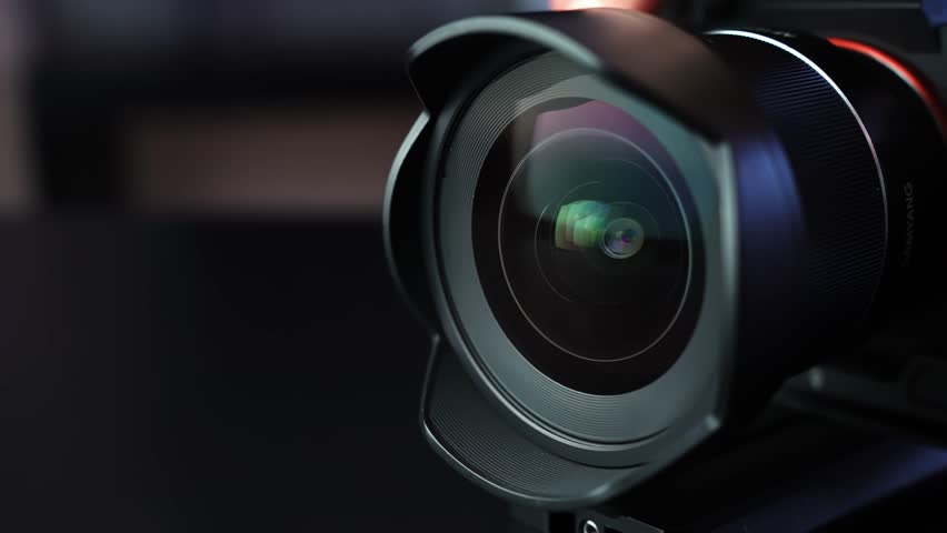 Cinematic macro of camera lens with hood as aperture opens wider, showing shifting glass reflections in dark studio lighting and shallow depth of field for tech b-roll.