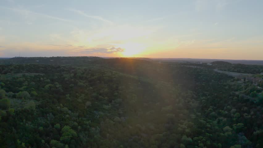 Aerial drone video flying west over trees into the setting sun. Blue skies with yellow-orange horizon around the sun.