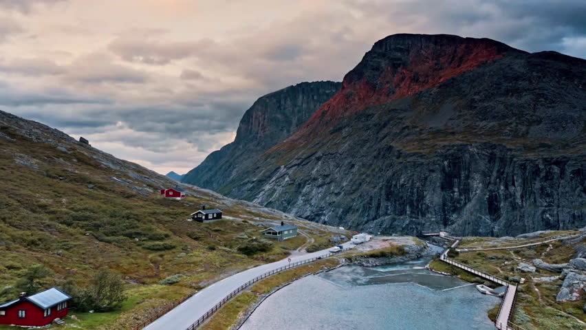 Aerial view of a serene mountain landscape featuring colorful cabins nestled among rocky terrain and lush greenery, with dramatic clouds overhead.