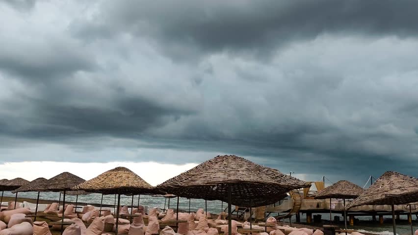 A cinematic wide shot of a quiet beach resort under a heavy, dramatic sky filled with dark storm clouds. The foreground features traditional straw sun umbrellas and empty bean bag chairs, suggesting a pause in summer activities. The grey sea meets the horizon under a moody atmosphere, capturing the raw beauty of nature before a rainfall. Perfect for projects focusing on climate, seasonal changes, travel cancellations, moody landscapes, or the peaceful solitude of an empty coastline.
