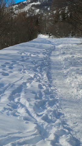 Personal perspective of walking along a narrow, snowy path in a winter mountain landscape. Fresh snow with footprints under a clear blue sky
