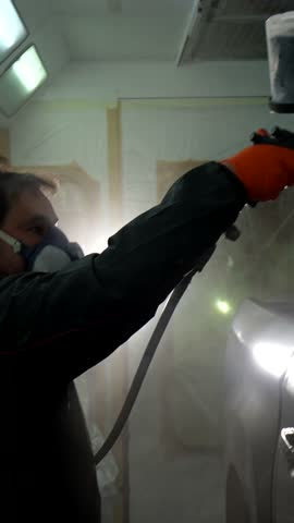 Automotive painter wearing a protective mask and gloves using an airbrush spray gun to paint a car part, working with precision inside a professional body shop spray booth with bright lights