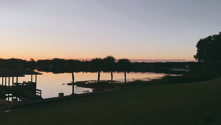 A calm lake stretches across the frame as the sun sets behind a distant tree line, casting a deep orange glow along the horizon. The sky transitions from warm amber near the water to soft muted tones above. A tall palm tree stands in silhouette on the left, its fronds sharply outlined against the fading light. On the right, a covered wooden dock extends over the water, its roof and railings forming clean geometric shapes mirrored clearly in the smooth surface below. The lake appears almost glasslike, reflecting the dark shoreline and glowing sky with near perfect symmetry. The foreground lawn remains in shadow, enhancing the contrast between the illuminated horizon and the silhouetted structures, creating a balanced and peaceful lakeside composition defined by stillness and warm evening color.