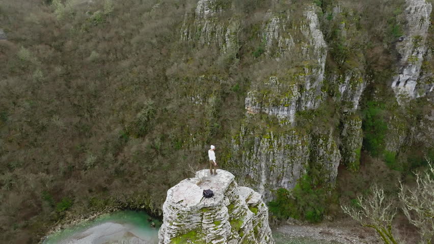 A lone man stands atop a stone formation in the rugged landscape of Zagorochoria under cloudy winter skies